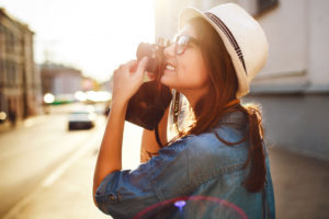 Outdoor summer smiling lifestyle portrait of pretty young woman having fun in the city in Europe in evening with camera travel photo of photographer Making pictures in hipster style glasses and hat