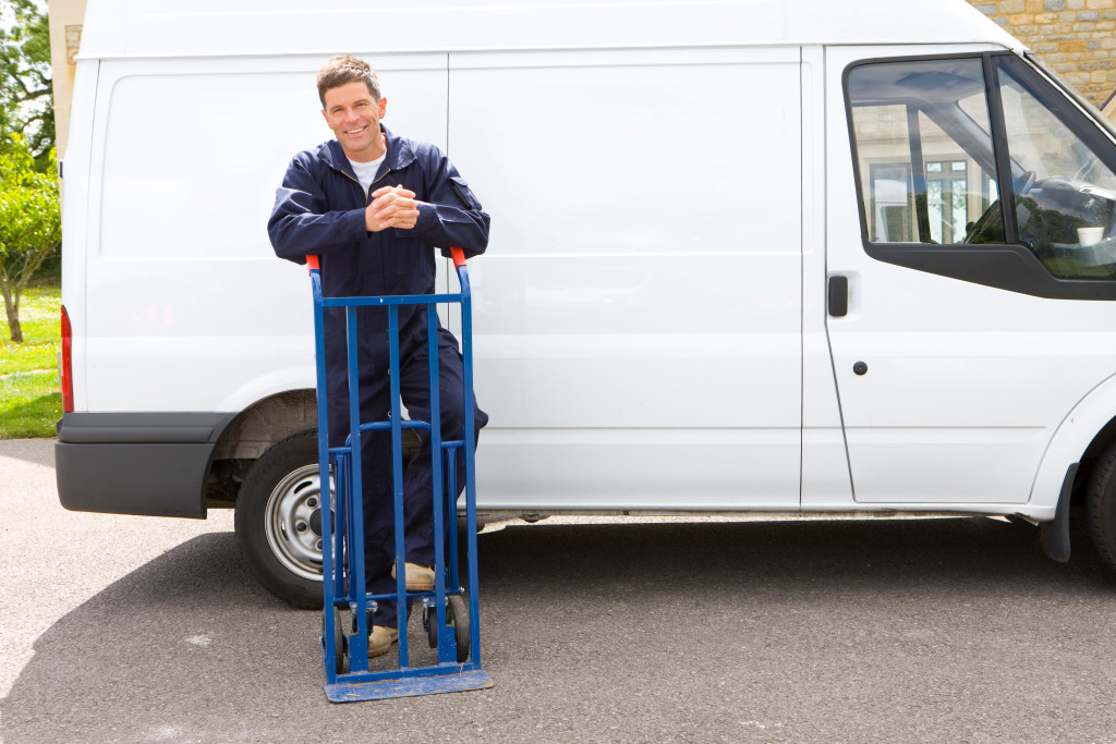 A man in coveralls smiling and leaning on a hand truck with a white work van