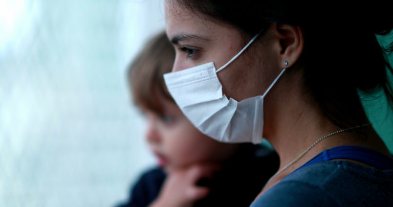 Mother wearing covid-19 mask holding baby looking outside standing by window