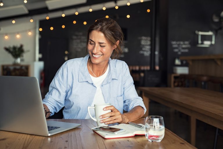 A freelance writer working in a cafe while holding a cup of coffee with a laptop and a journal on her table