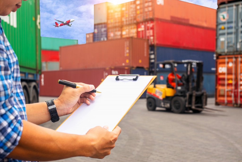 a man holding a clipboard with a plane, a forklift, and a bunch of shipping containers in the background