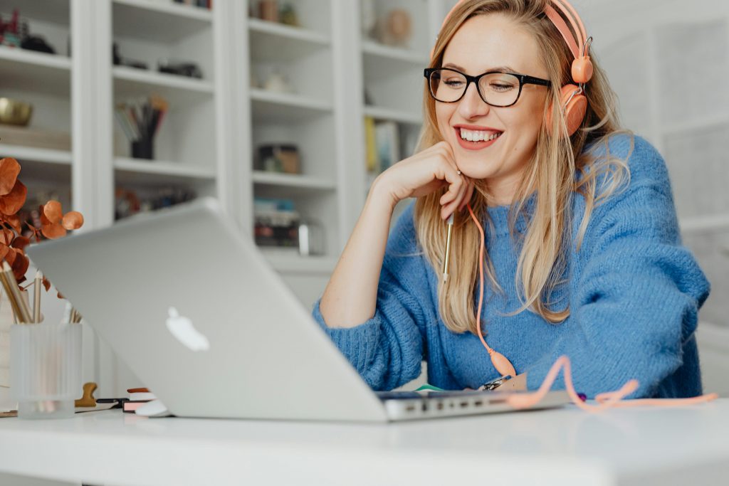 woman in a blue sweater and pink headphones smiling while working on a laptop
