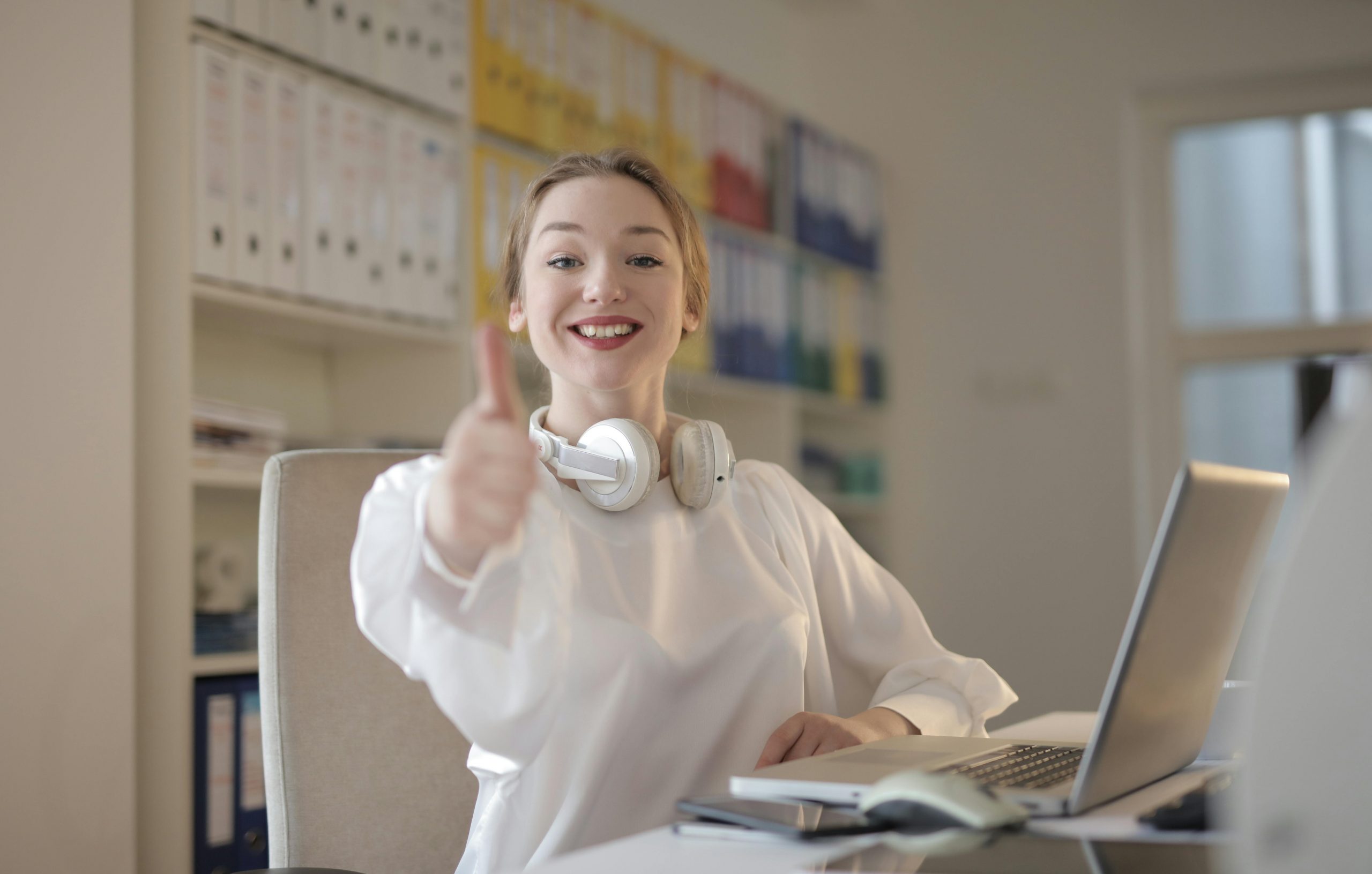 smiling woman giving a thumbs-up, with headphones resting on her shoulders at her home desk